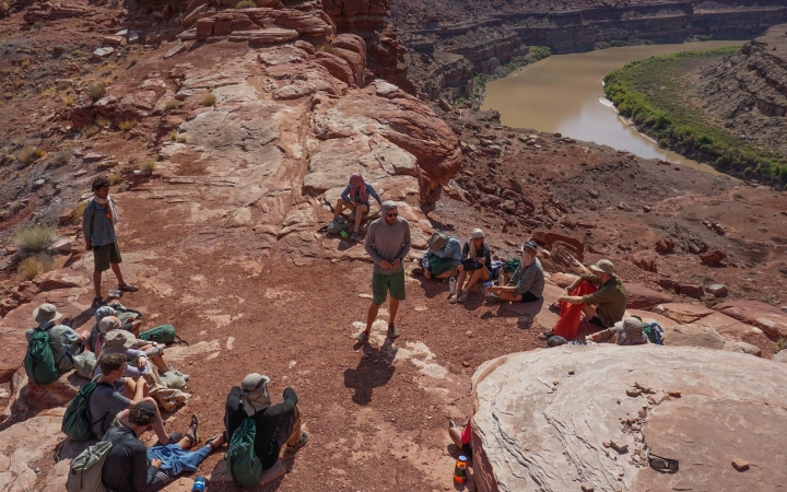 A group of students sit on the ground while listening to an instructor provide information. Below them, a river winds through canyon walls.